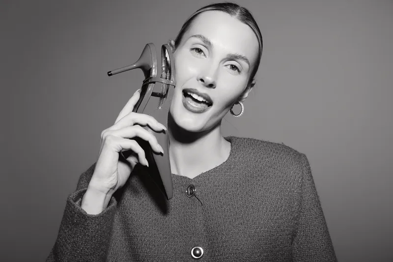 Caucasian woman with pale skin holds high heel shoe near face, wearing cozy sweater and hoop earrings in studio on grey background.