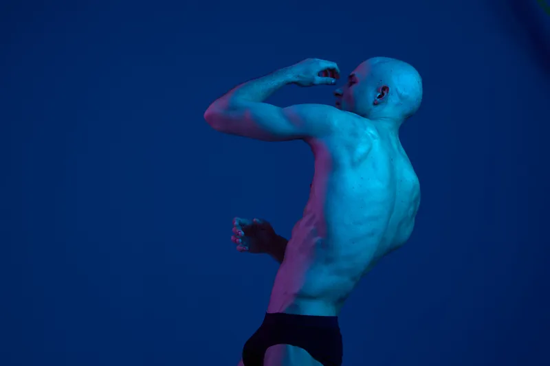 Athletic man posing dramatically with blue lighting, showcasing strength and movement in a captivating studio setting. Emphasizes physical fitness and artistry.