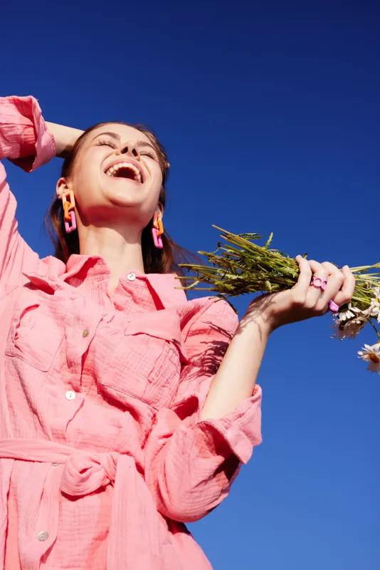 Joyful young woman in pink dress laughing while holding wildflowers against a bright blue sky, embodying happiness and nature in spring