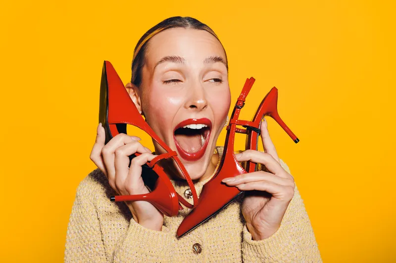 Happy woman with red high heels holds shoes near face, smiling with open mouth on bright yellow background. Concept for fashion, style, shopping, and joyful emotions.
