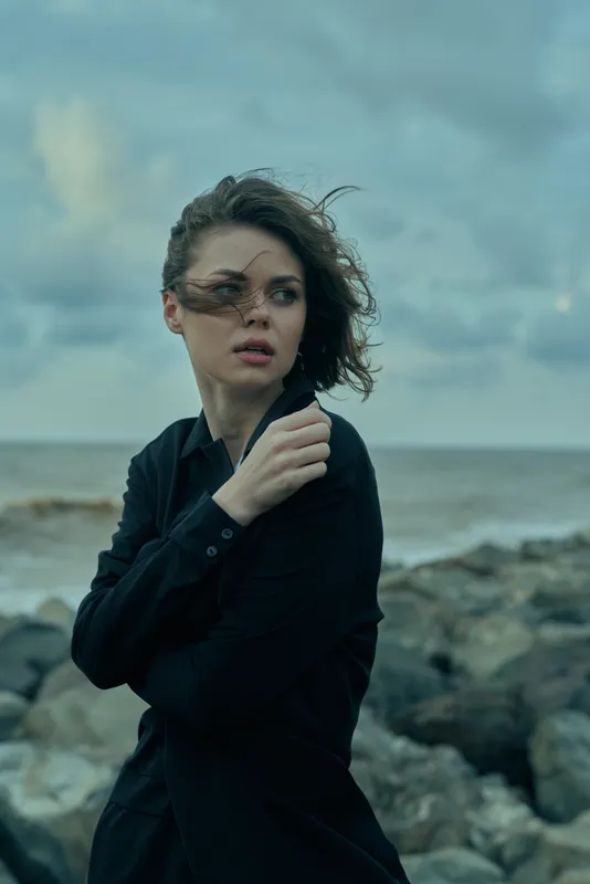 Windy coastal scene with a woman in a dark jacket near a rocky shoreline, hair and clothing blown by gusts, moody atmosphere and a distant horizon