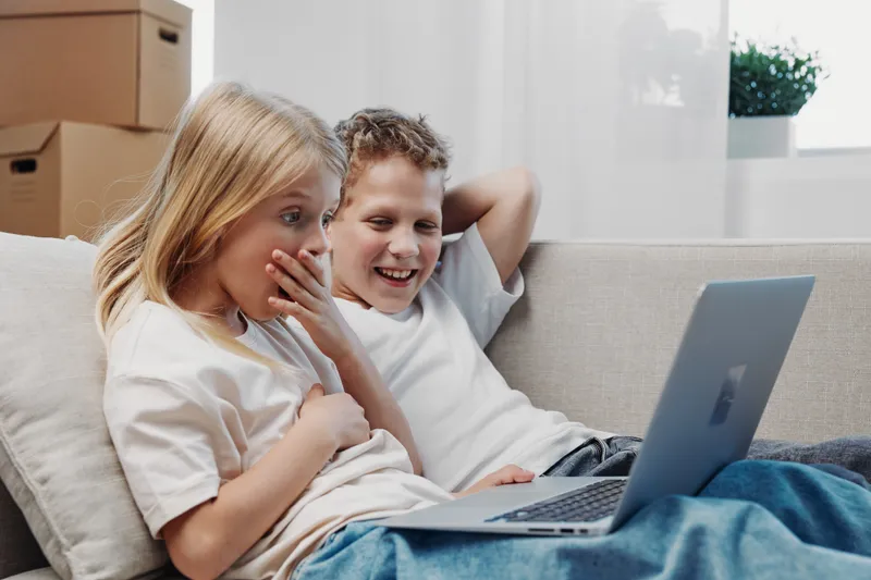 Two children in white shirts sitting on a couch watching laptop, showing surprise and laughter, casual home environment with natural light and moving boxes behind. Concept for family leisure and
