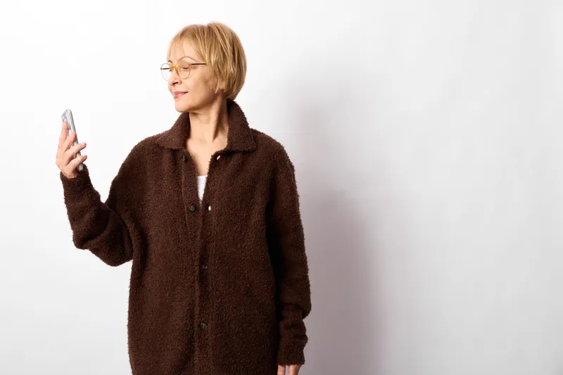Woman smartphone annoyance stress communication technology portrait indoors studio texting. Mature woman in brown coat holds a phone, tense, annoyed by communication on white background.
