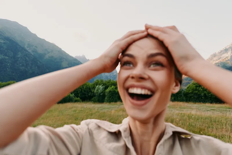 Young woman outdoors smiles broadly, hands on head in a sunny meadow with mountains behind, natural light, fresh air, carefree vibe.