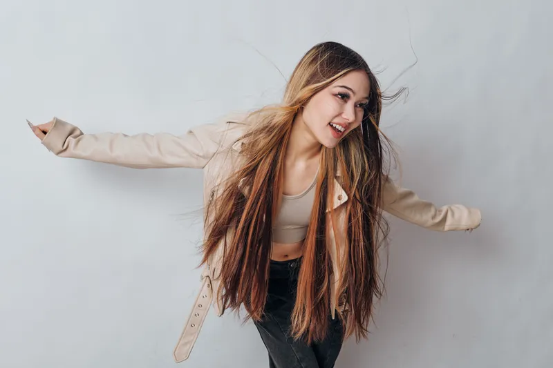 Young woman with long hair wearing beige jacket and crop top dancing or posing against white background with casual expression and movement.