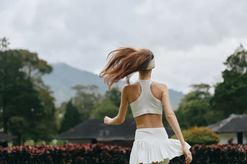 Motivated young woman running in a white sports outfit, showcasing athleticism and vitality, set against a lush green landscape under a cloudy sky