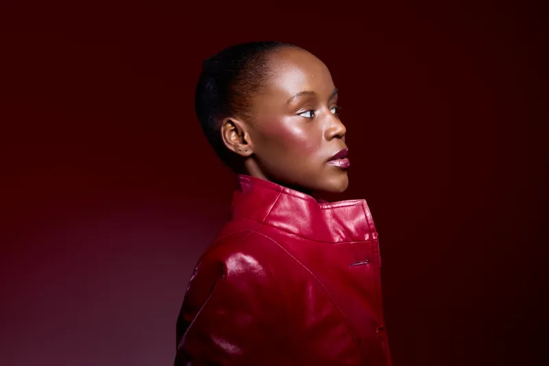 Profile portrait of African American woman with dark skin wearing red leather jacket on dark red background. Concept of fashion, beauty, confidence, and elegance in studio lighting.