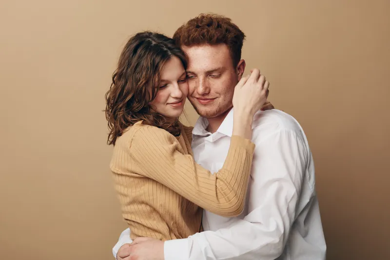 Young couple embraces tenderly with closed eyes, expressing love and affection. Warm beige background and casual clothing create intimate and cozy atmosphere.