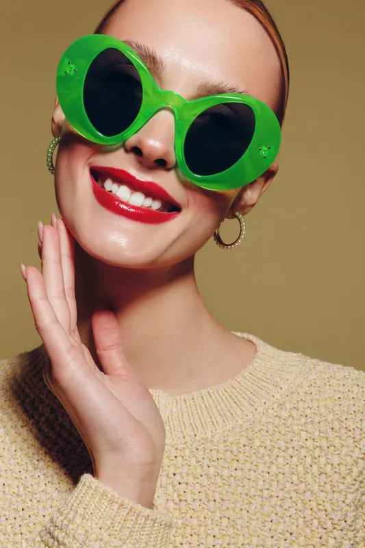 Smiling woman with bright red lipstick wears oversized green sunglasses and beige sweater. Close-up portrait with hoop earrings on neutral background. Concept of fashion, style, and confidence.