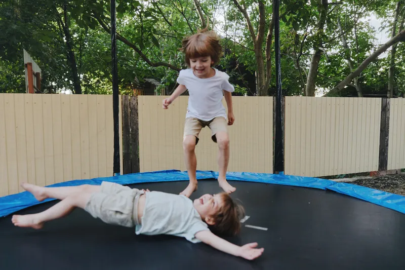 Joyful children playing on a trampoline, one boy in white shirt, smiling as he jumps, surrounded by greenery and a sunny atmosphere.