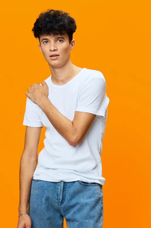 young man wearing white t-shirt and jeans posing thoughtfully with curly hair against bright orange background, casual style and relaxed mood