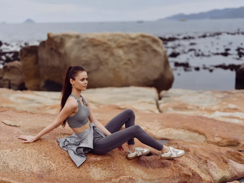 Stylish young woman in activewear sitting on rocks by the water, exuding confidence and relaxation, embodying fitness and tranquility