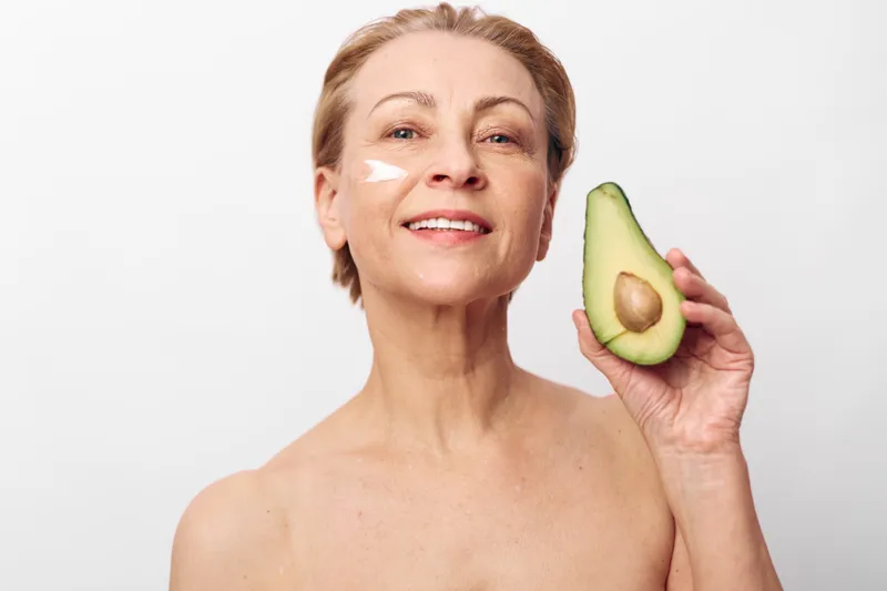 Smiling mature woman with natural skin holds half avocado near face, applying white cream on cheek. Concept of healthy skincare, natural beauty, organic wellness, and self-care routine.