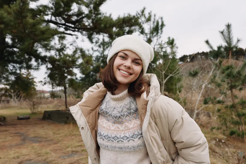 Smiling woman in a warm beige coat and knit sweater outdoors in a wintry park, posing warmly with a casual, candid style.
