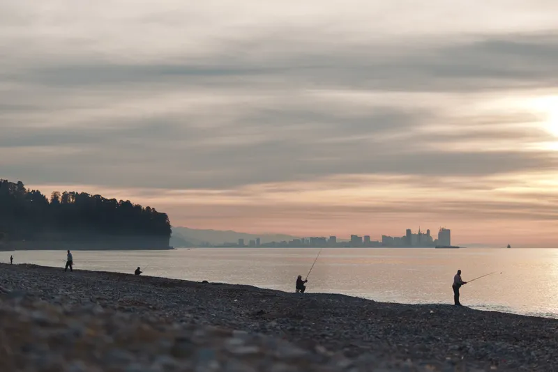 Beach at sunset with silhouettes of anglers along a pebbled shore, calm water and distant city skyline, soft clouds and warm light create a tranquil coastal scene for leisure and reflection.