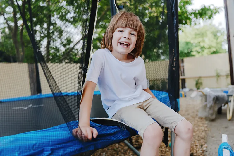 Happy boy enjoying a sunny day, smiling while sitting on a trampoline, surrounded by greenery and bright outdoor colors for a joyful atmosphere.