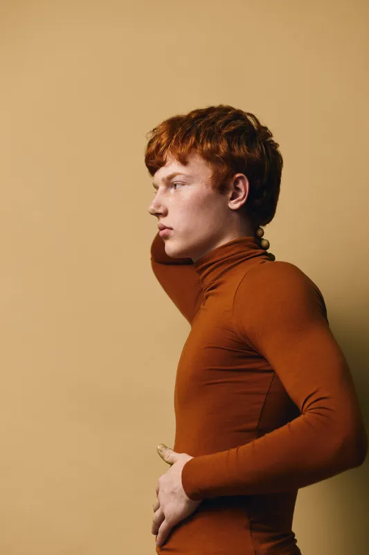 Young man with red hair wearing brown turtleneck posing in profile against beige background with thoughtful expression and hand behind neck for casual portrait.
