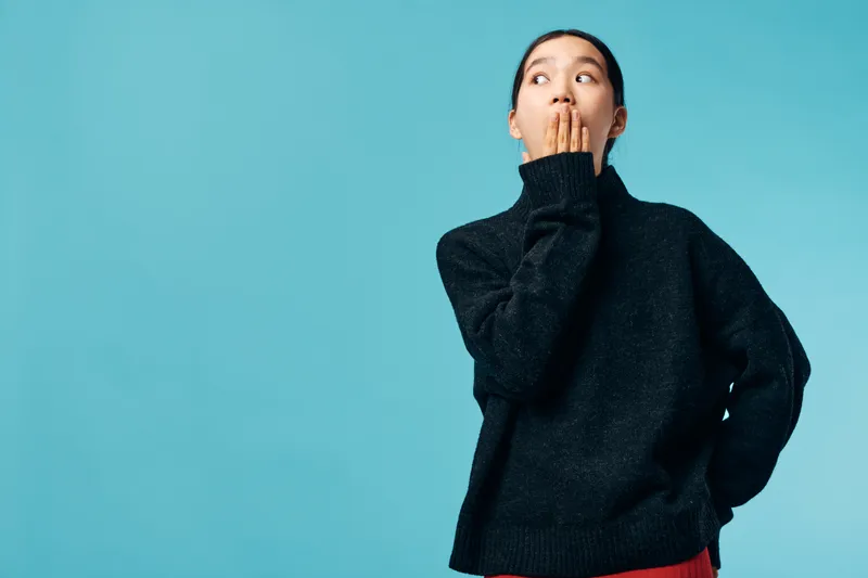 Young woman with hand near mouth showing surprise or shock expression standing against blue background wearing black sweater with casual pose and thoughtful look.