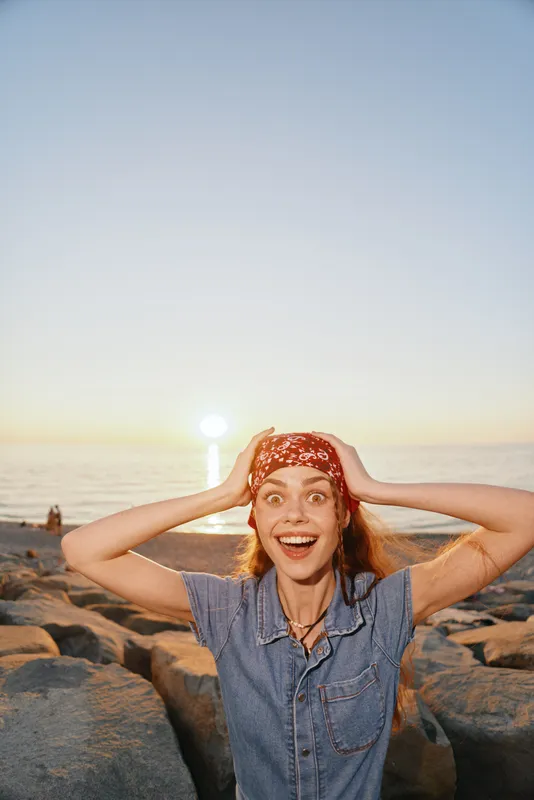 happy woman in denim shirt and red headband enjoying sunset at rocky beach with clear sky, natural light, and warm summer atmosphere