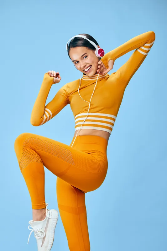 Fitness woman in vibrant orange athletic outfit performs dynamic pose against blue backdrop. Sporty wear, headphones, and energetic movement highlight cardio training, dance-inspired workout,