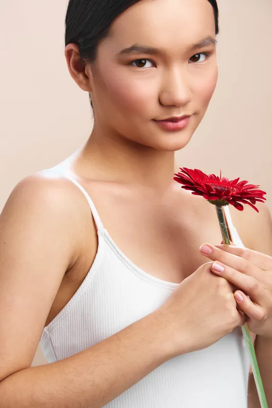 asian woman holds a red flower near her chest, wearing a white tank top, serene expression, soft lighting, intimate portrait conveying gentleness and beauty