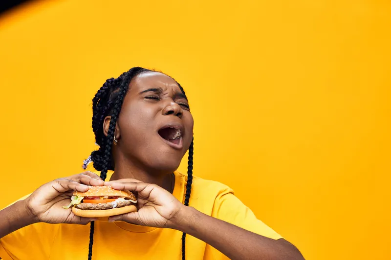 Happy girl enjoying a delicious burger against a vibrant yellow background, showcasing the joy of eating fast food and healthy lifestyle choices
