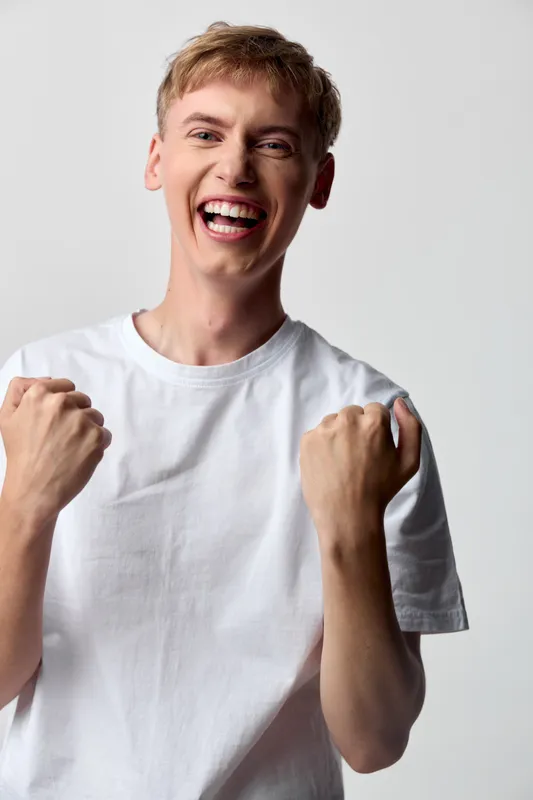 Joyful confident young man with white tee raises fists in celebration, bright smile and upbeat energy, studio style, casual vibe, conveying motivation, success mindset and cheerful determination