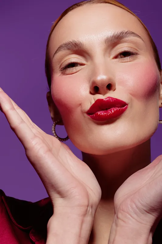Close-up of a woman with glossy red lipstick blowing a kiss, wearing hoop earrings and a burgundy top. Purple background highlights her smooth skin and confident expression.