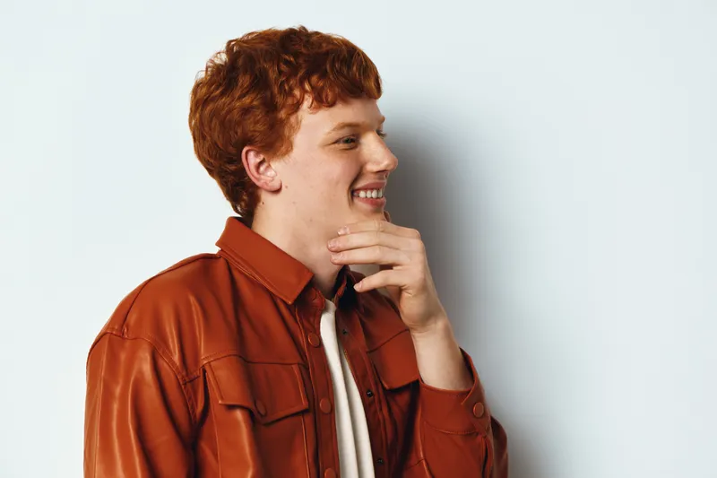 Young man with red hair and smile dressed in orange jacket and white shirt looking to side and touching chin against plain background with positive expression.