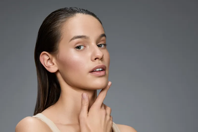 Young woman with natural beauty and clear skin, showcasing skincare routine and healthy lifestyle, in neutral tones against a soft gray background