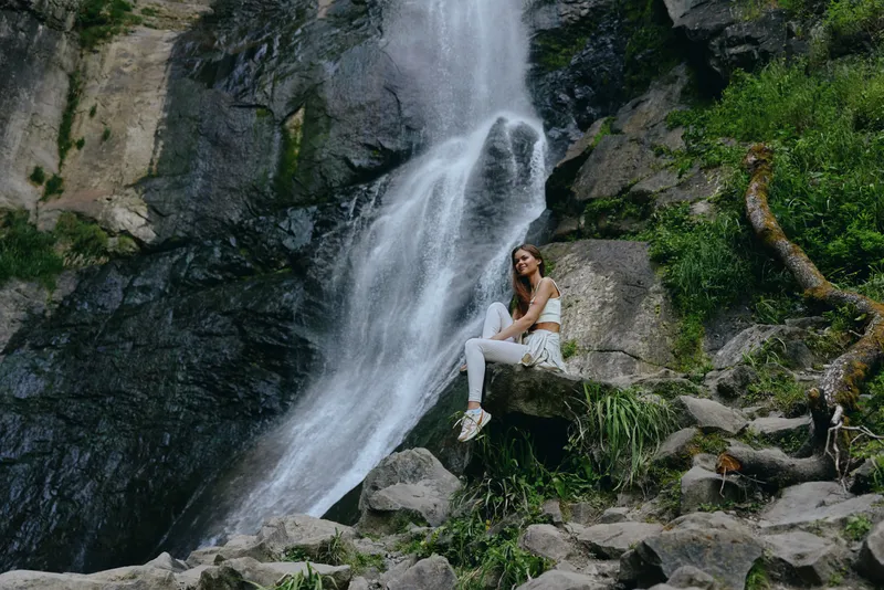 Young woman sitting on rock near waterfall in lush green forest, wearing white clothes and smiling peacefully in natural outdoor setting.