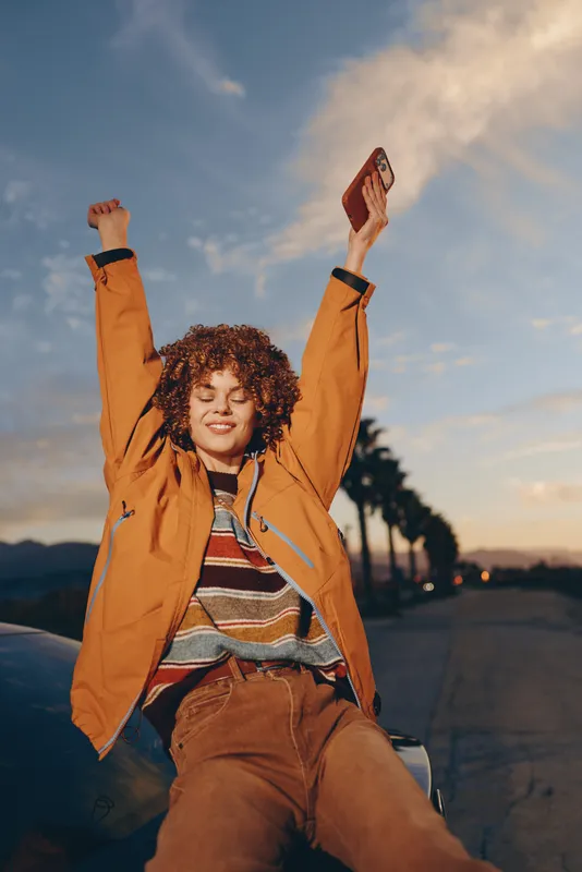 Woman curly hair outdoors lifestyle sunset sky joyful arms casual jacket pose, smiling while sitting on a pavement walkway at dusk, golden hour light, relaxed carefree travel mood