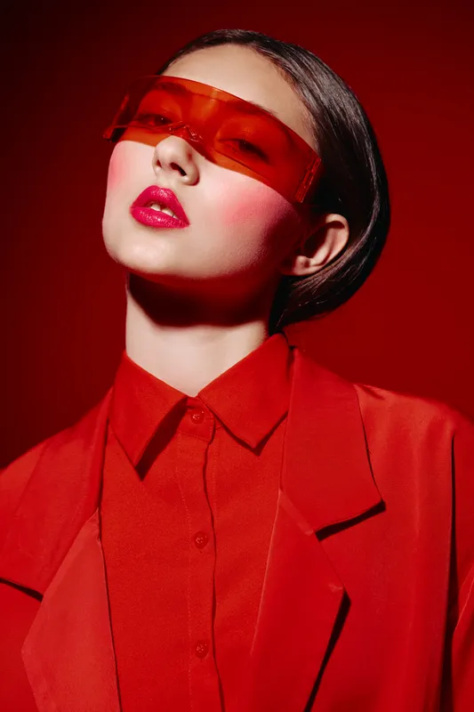 Young woman in red outfit wears futuristic orange visor and vibrant makeup. High fashion creative portrait in studio lighting on bold red background. Concept of beauty, style.