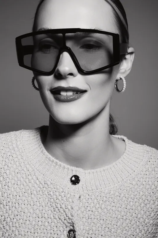 A Caucasian woman with fair skin wears oversized futuristic sunglasses and a knitted sweater. Studio portrait in black and white with modern fashion and style concept.