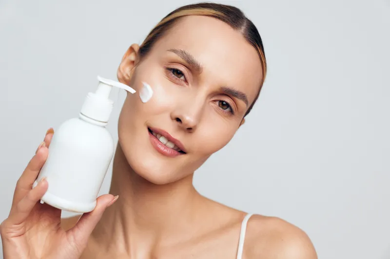 Young woman with clear skin holding white lotion bottle applying cream on cheek. Close-up on light background. Concept of skincare, beauty, health, and self-care routine.