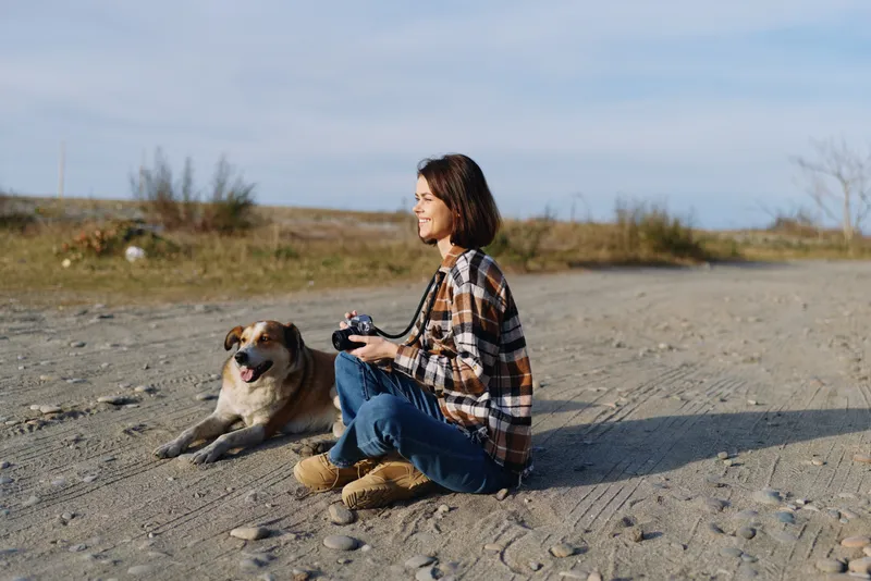 Woman sitting on sandy ground with her dog in an open desert landscape, holding a camera remote, enjoying quiet outdoor moments and companionship