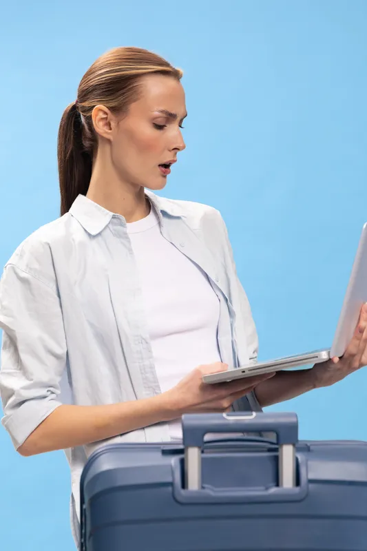 Surprised woman with laptop stands behind blue suitcase on blue background. Concept of travel planning, unexpected news, and business trip preparation.