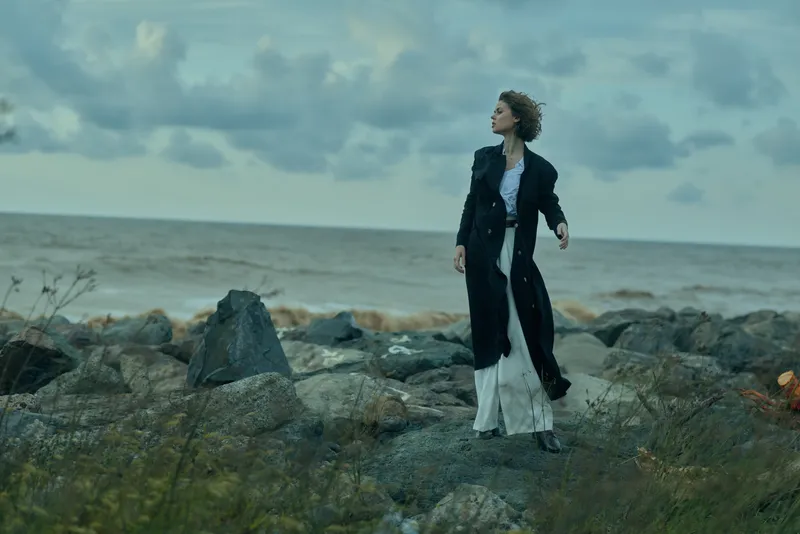 Woman on a rocky shoreline in a long dark coat gazing at the sea under a moody sky, blending fashion with rugged nature and a sense of solitude