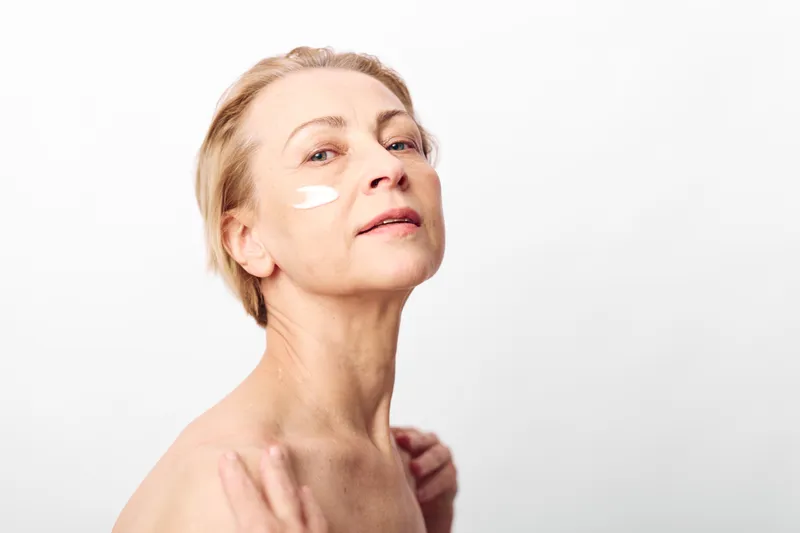 Mature woman with short blond hair applies white face cream on cheek, looking confident against white background. Concept of skincare, aging, beauty routine, and self-care for older skin.