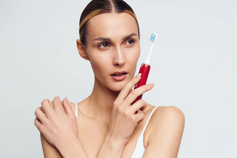 Woman applying skincare to face in studio, holding toothbrush and red serum bottle. Minimal white background, soft studio lighting, calm mood, beauty, wellness, selfcare.