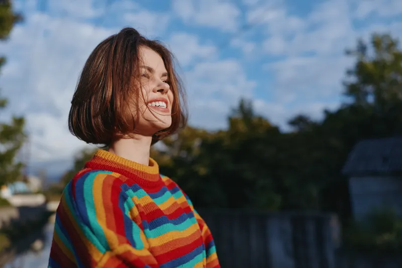 Woman smiling young adult in striped sweater outdoor portrait, a cheerful 20s brunette with bob haircut leaning into sunlight, casual outfit for colorful travel lifestyle and cozy fashion mood.