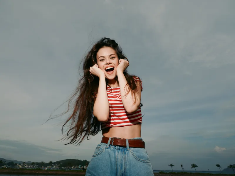 Joyful young woman outdoors wearing a striped crop top and denim jeans, hands on cheeks, wind-swept hair, sunny sky, carefree mood and vibrant energy.