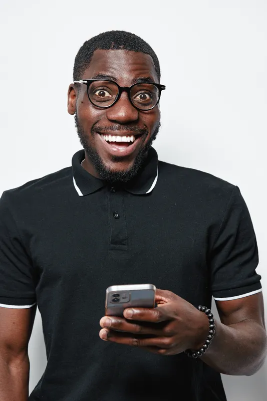African American man with dark skin wearing glasses and black polo shirt smiling while holding smartphone isolated on white background in studio portrait.