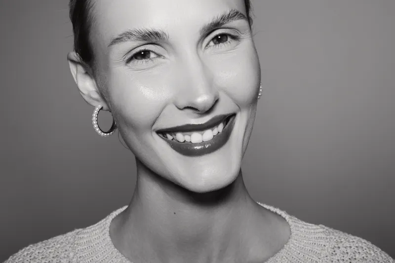 Portrait of a smiling Caucasian woman with fair skin and short hair, wearing hoop earrings and a knitted sweater in a studio setting with soft lighting.