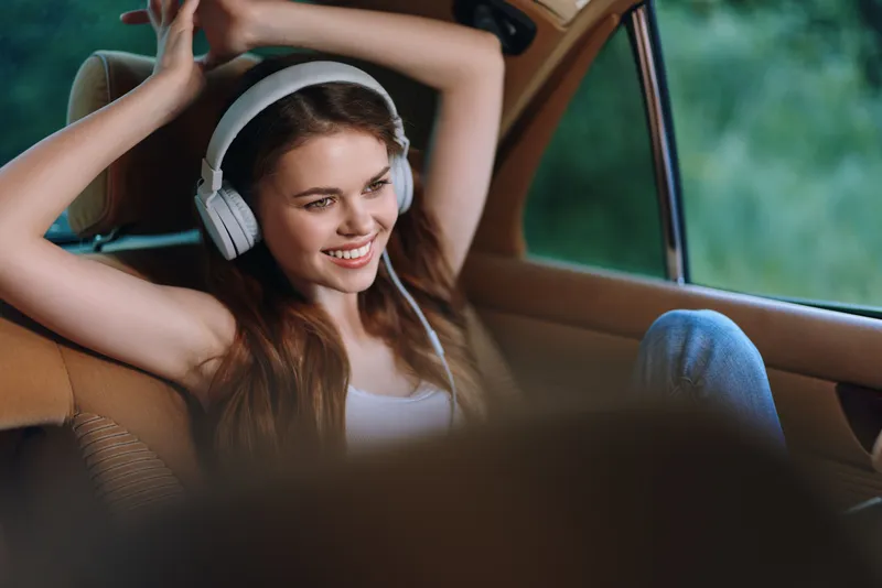 Young woman smiling with headphones in a car, enjoying music and relaxation in a natural green background, perfect for lifestyle and travel themes.