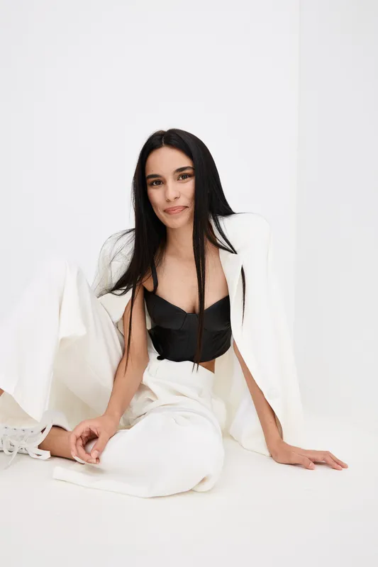 Young woman with long dark hair sitting on the floor dressed in white pants and jacket with black top, smiling softly in a bright minimalistic studio setting.