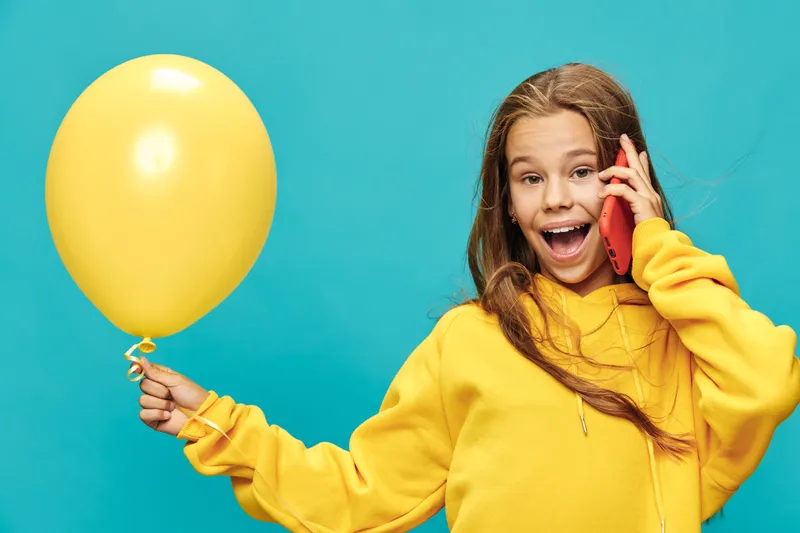 Girl in yellow hoodie holding balloon and talking on phone with excited expression on blue background. Child, communication, celebration, happiness, casual clothing, cheerful mood.