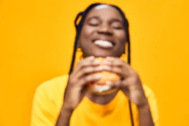 Happy young woman enjoying a burger on a vibrant yellow background, expressing joy and satisfaction, highlighting the concept of healthy eating and food enjoyment