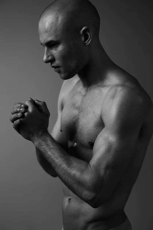 Thoughtful man posing in black and white, showcasing strength and emotion, highlights muscular physique and contemplative nature in a dramatic setting.