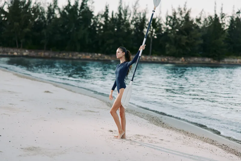 Young woman carrying paddle on beach wearing navy swimwear near tropical water with trees.
